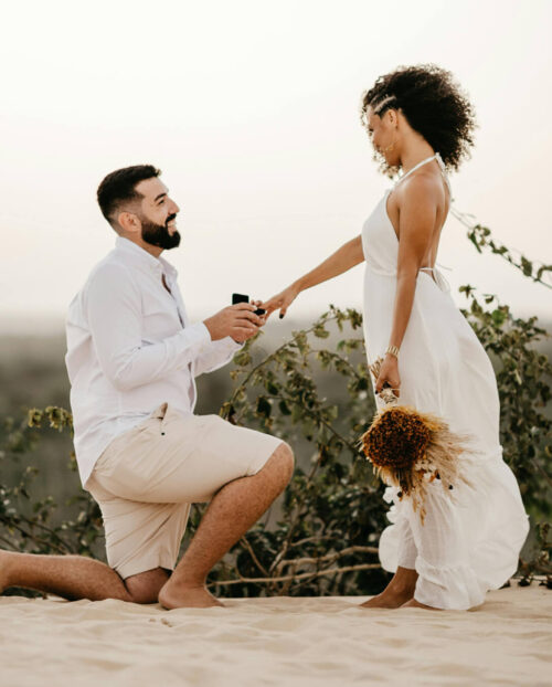 A man on one knee proposing to woman in a white dress