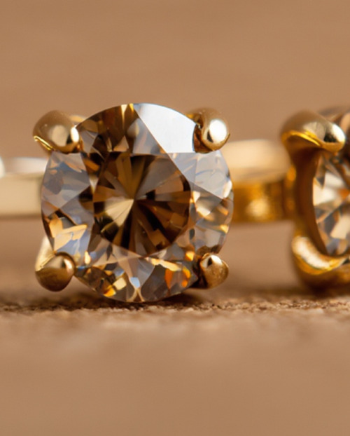 A pair of round Sunset Brown diamond stud earrings set in yellow gold, resting on a soft brown surface that highlights their warm tone and brilliance.
